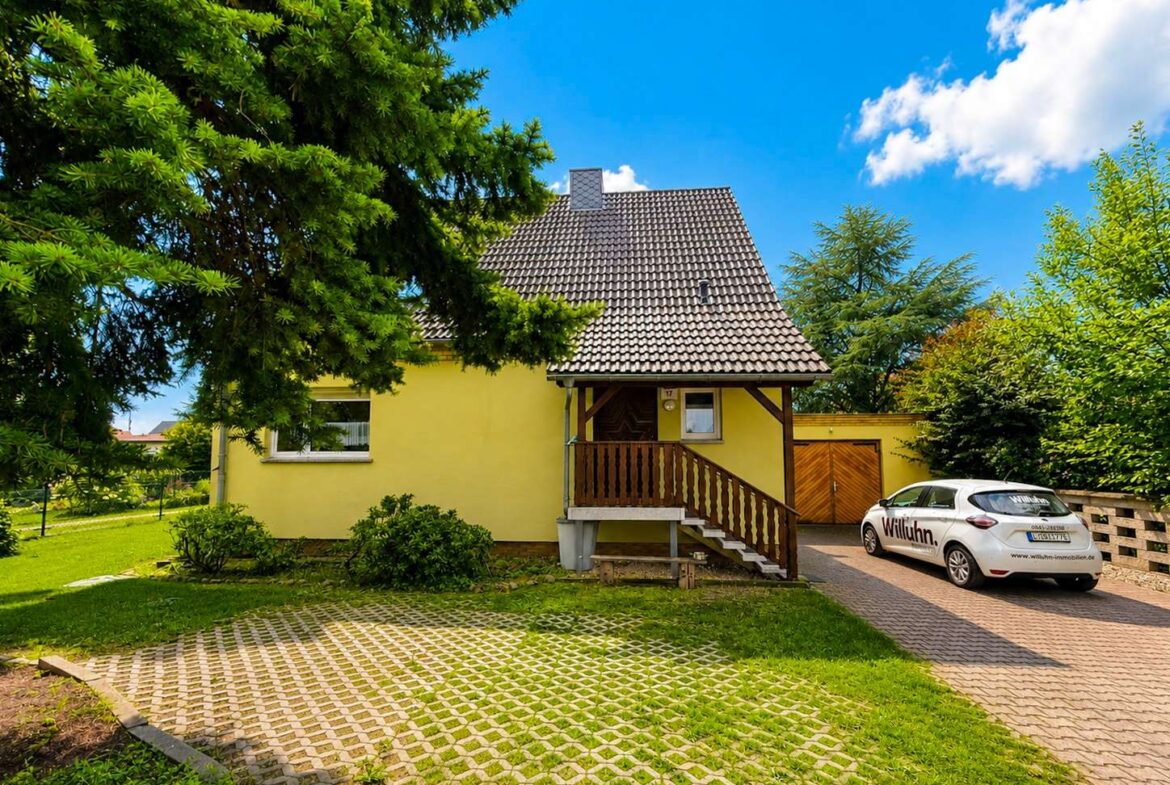 Yellow suburban house with brown-tiled steep roof, front porch, and wooden stairs, surrounded by green trees on a sunny day.