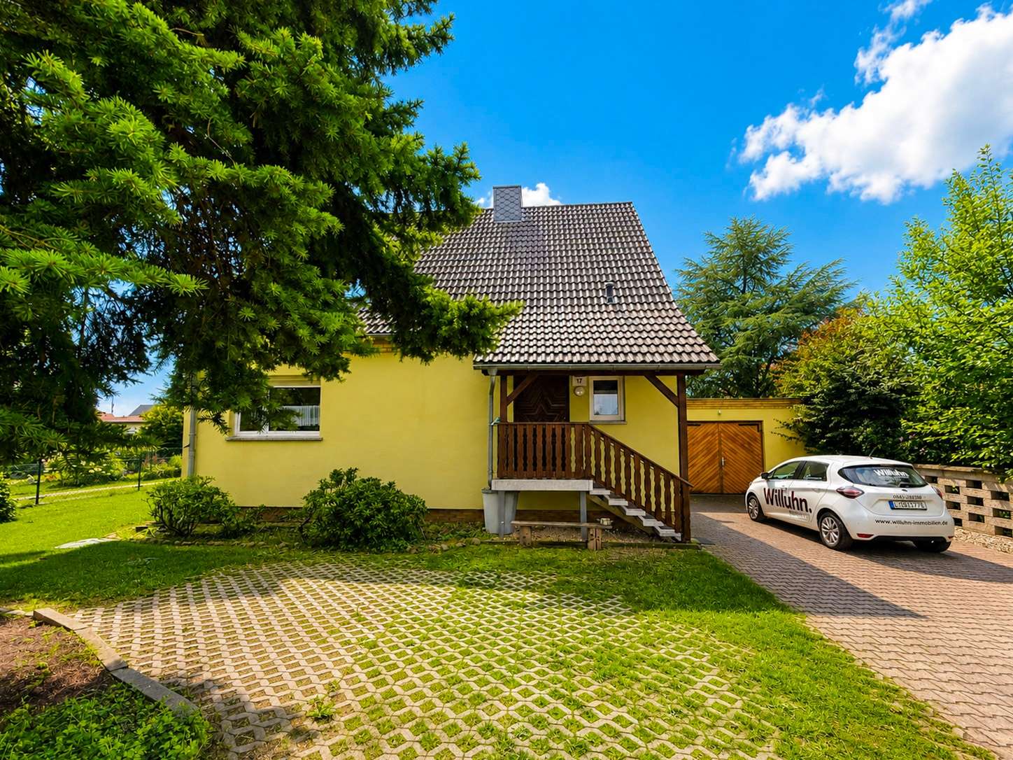 Yellow suburban house with brown-tiled steep roof, front porch, and wooden stairs, surrounded by green trees on a sunny day.