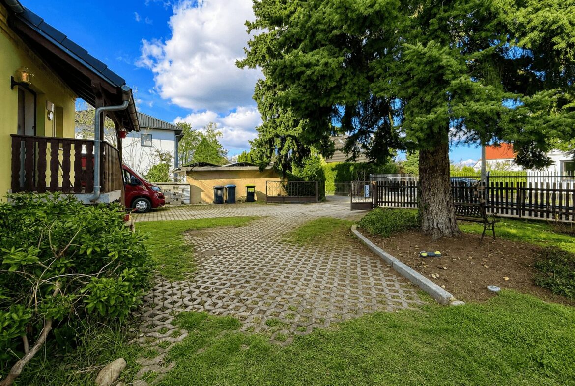 Yard of a yellow house with a wooden porch, a red car parked near the entrance, and a large tree to the right under a blue sky.
