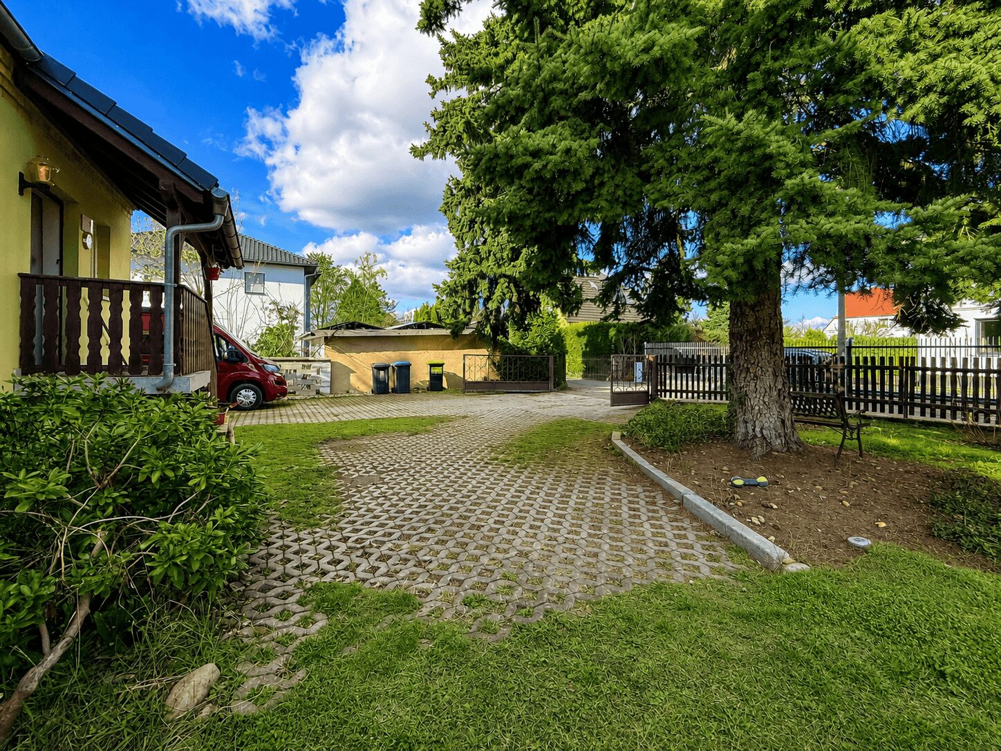 Yard of a yellow house with a wooden porch, a red car parked near the entrance, and a large tree to the right under a blue sky.