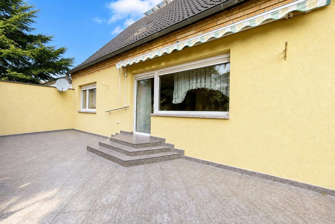 Yellow house exterior with a glass door, large window with lace curtains, and a tiled patio with a short brick stair set under a striped awning.