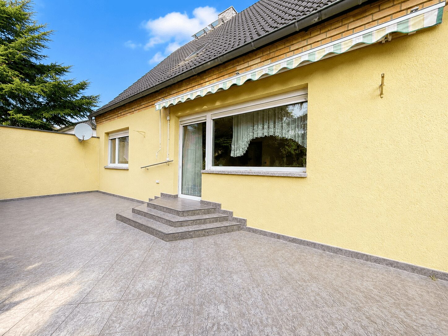 Yellow house exterior with a glass door, large window with lace curtains, and a tiled patio with a short brick stair set under a striped awning.