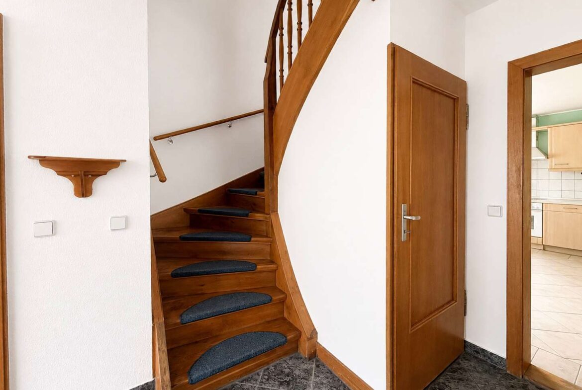 Wooden staircase with dark blue carpet runners, white walls, and a doorway to a kitchen on the right.