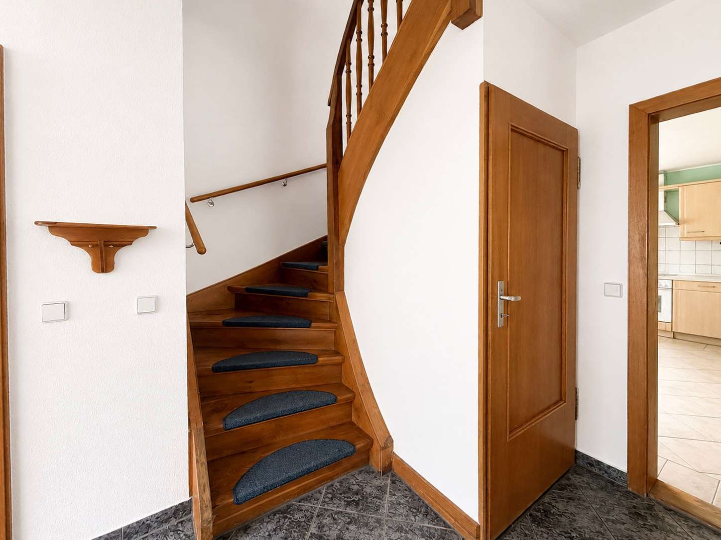 Wooden staircase with dark blue carpet runners, white walls, and a doorway to a kitchen on the right.