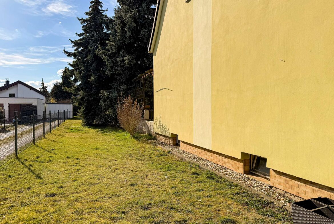 Yellow building with brick base on right, a grassy yard and chain-link fence along the left under a blue sky with trees in the background