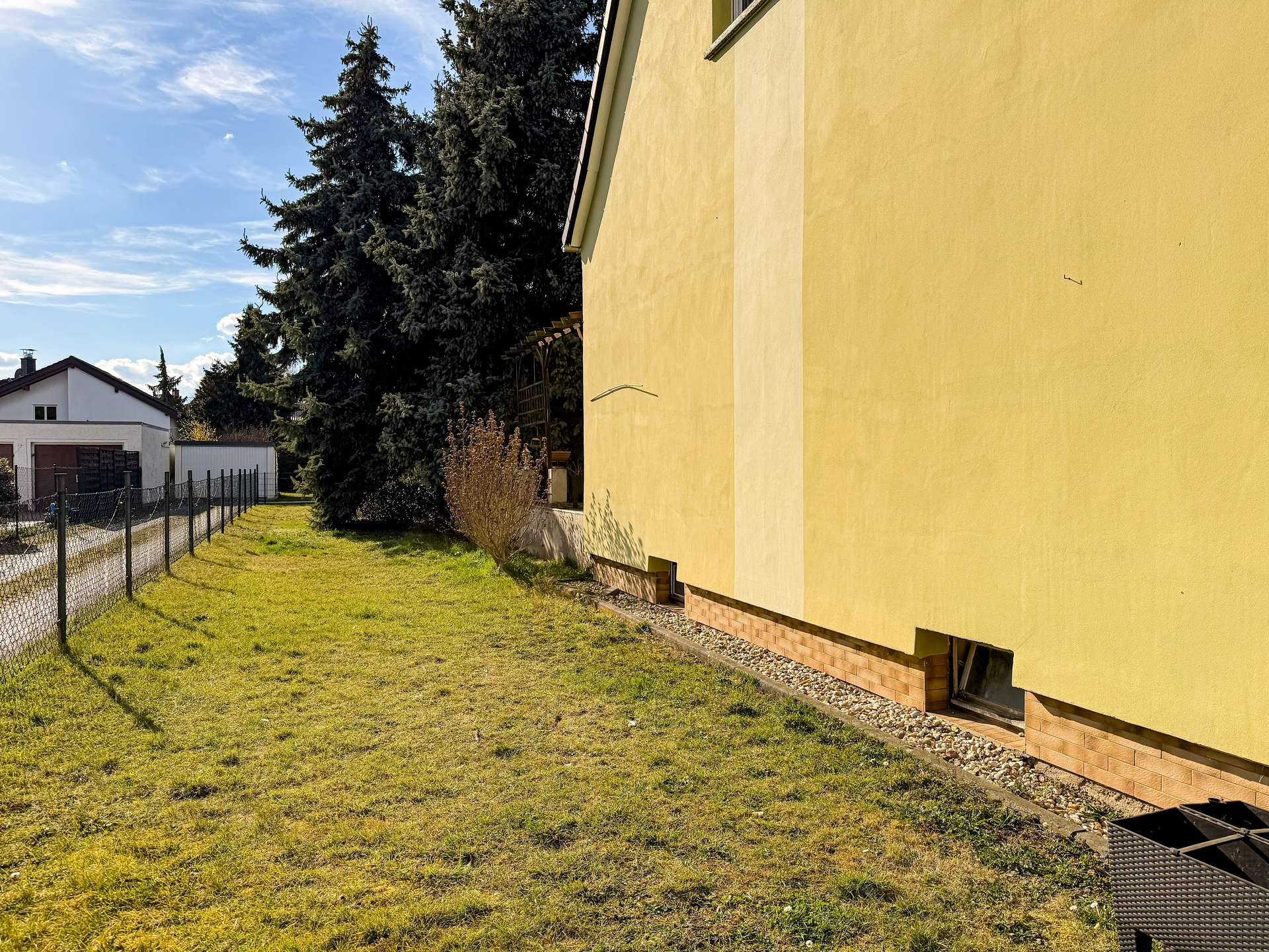 Yellow building with brick base on right, a grassy yard and chain-link fence along the left under a blue sky with trees in the background