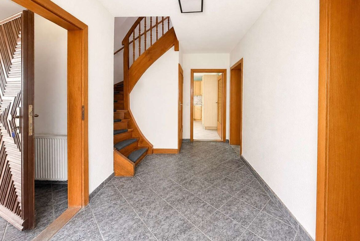 Bright hallway with gray tiled floor, white walls, and a curved wooden staircase on the left.