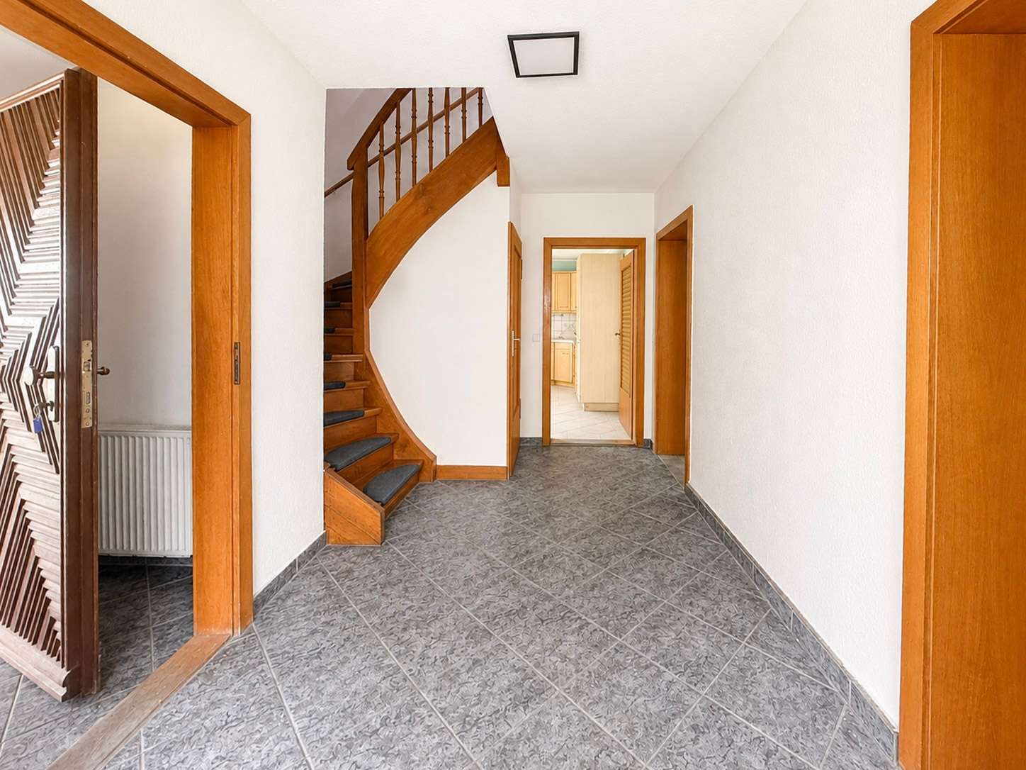Bright hallway with gray tiled floor, white walls, and a curved wooden staircase on the left.