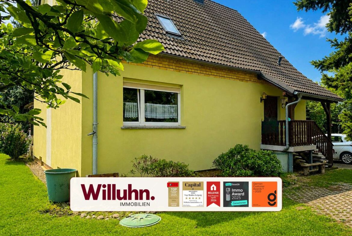 Yellow house with brown tiled roof and a front lawn; a large Willuhn Immobilien banner with awards in the foreground.