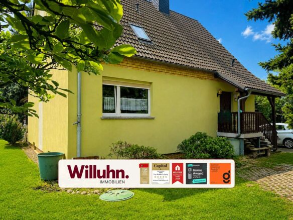 Yellow house with brown tiled roof and a front lawn; a large Willuhn Immobilien banner with awards in the foreground.