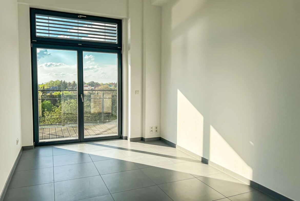 Bright empty room with a large glass door to a balcony, daylight casting diagonal shadows on the gray tiled floor.