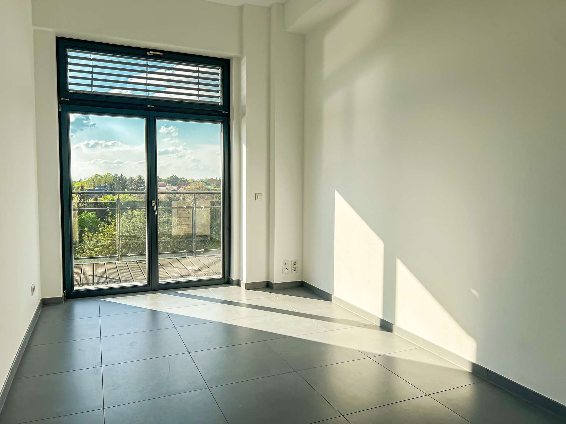 Bright empty room with a large glass door to a balcony, daylight casting diagonal shadows on the gray tiled floor.