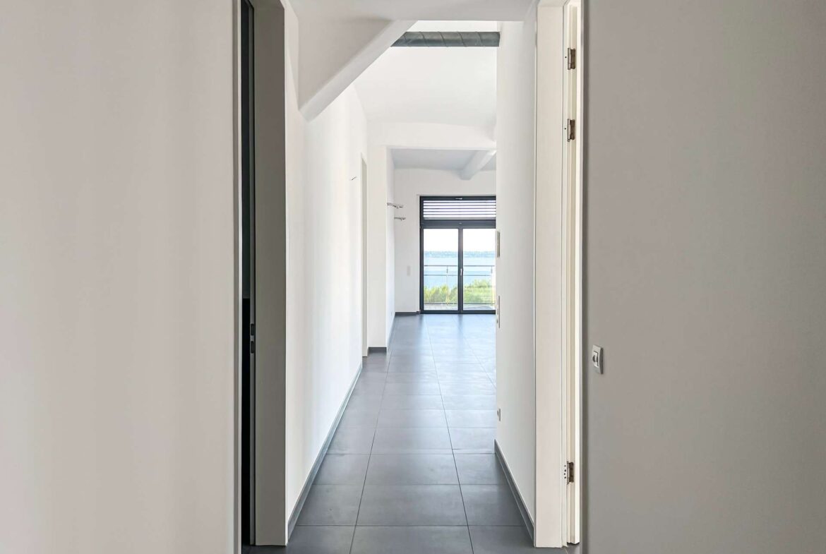 Bright hallway with white walls and grey tiled floor, ending at a glass door with an ocean view outside