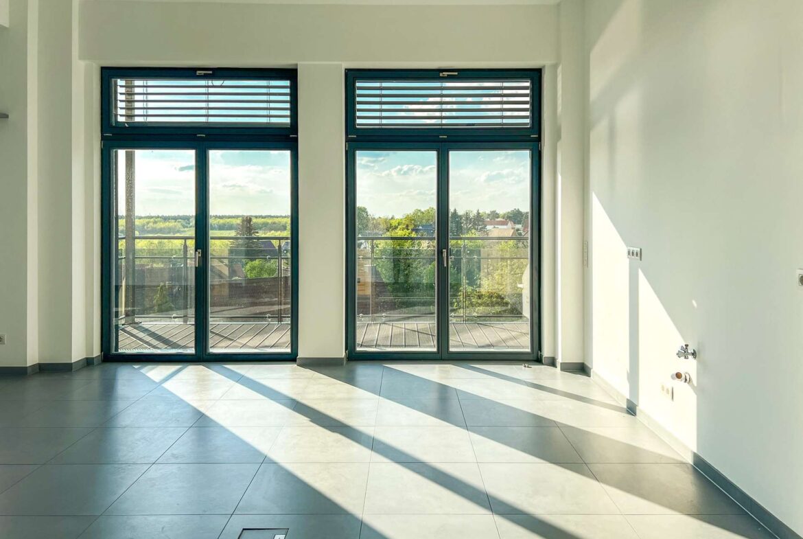 Modern empty room with large blue-framed glass doors opening to a balcony and a view of trees outside.