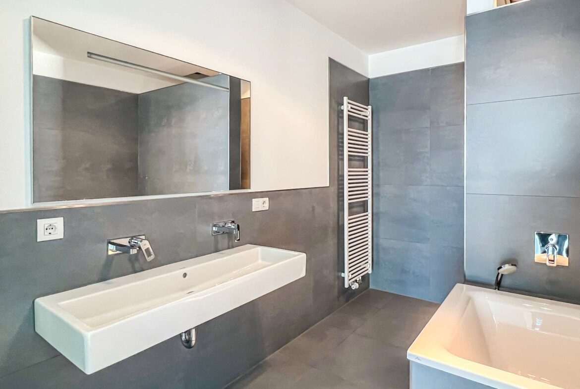 Modern bathroom with long rectangular white sink, gray tiled walls, and a wall-mounted mirror.