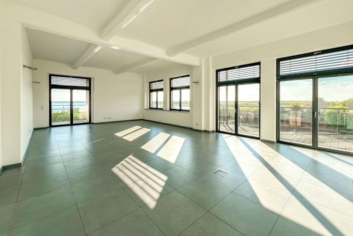 Bright empty living room with white walls, exposed beams, and large glass doors to a balcony overlooking water.