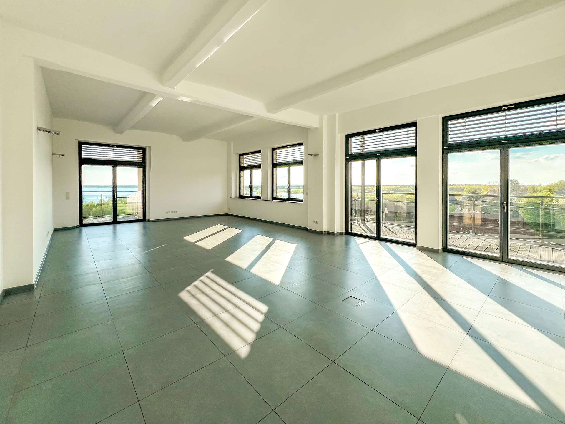Bright empty living room with white walls, exposed beams, and large glass doors to a balcony overlooking water.