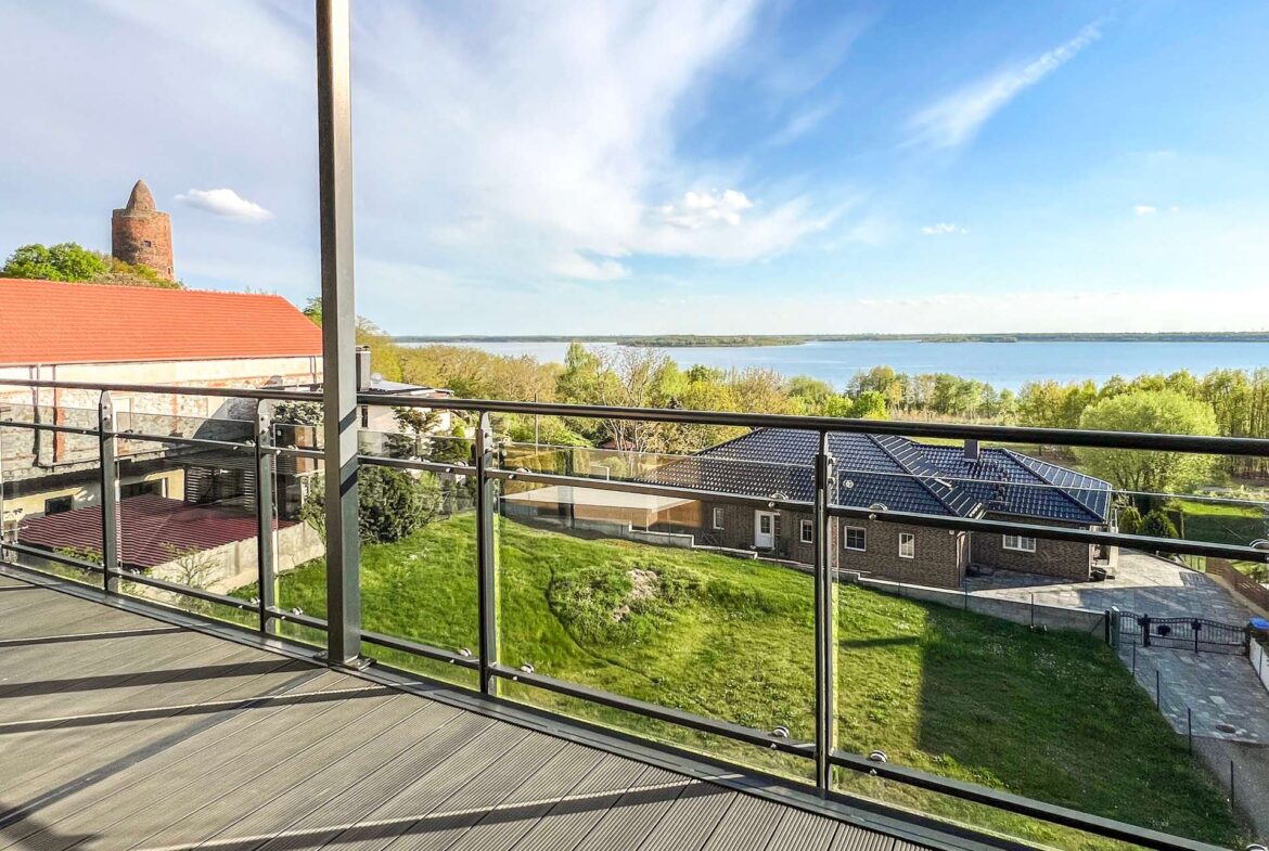 Balcony with glass railing overlooking a green lawn, red-roofed buildings, and a calm lake under a blue sky.