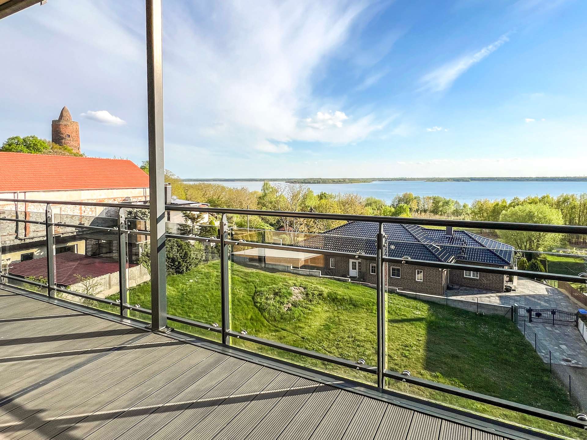Balcony with glass railing overlooking a green lawn, red-roofed buildings, and a calm lake under a blue sky.
