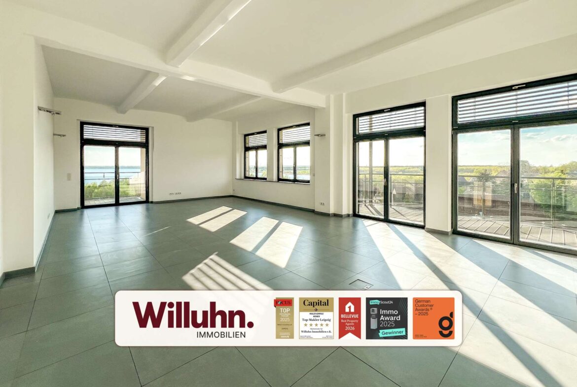 Bright empty living room with large windows and balcony doors; view of trees and water outside; Willuhn Immobilien banner at bottom.