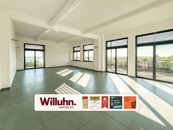 Bright empty living room with large windows and balcony doors; view of trees and water outside; Willuhn Immobilien banner at bottom.