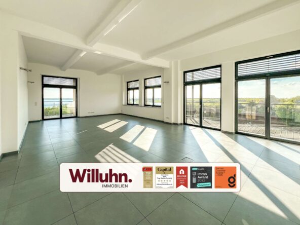 Bright empty living room with large windows and balcony doors; view of trees and water outside; Willuhn Immobilien banner at bottom.