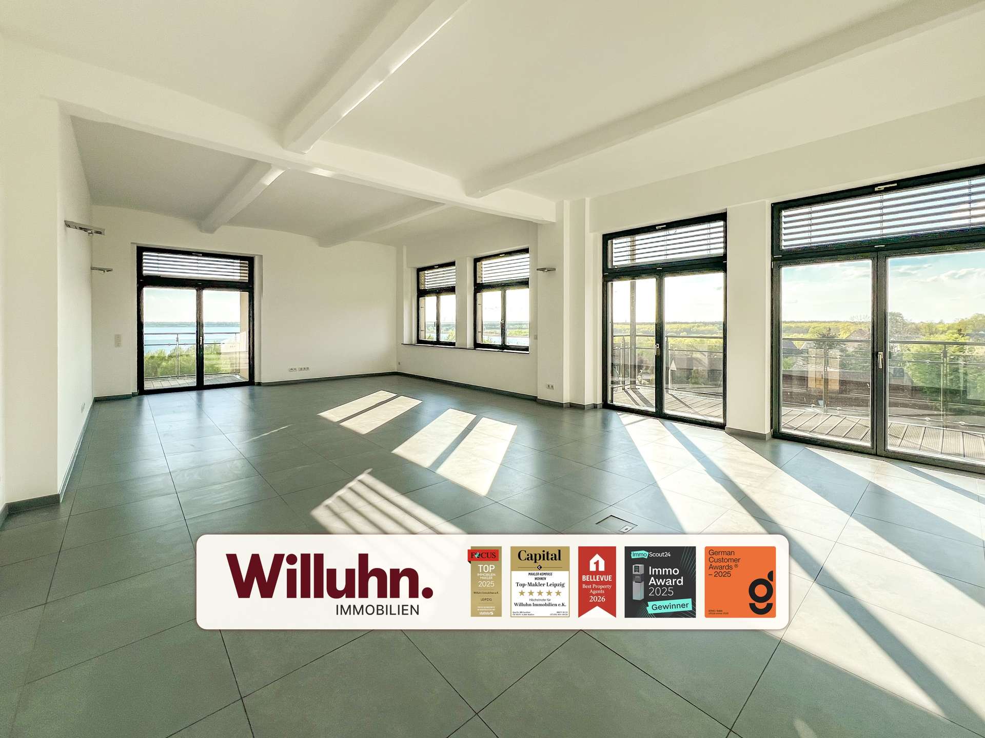 Bright empty living room with large windows and balcony doors; view of trees and water outside; Willuhn Immobilien banner at bottom.