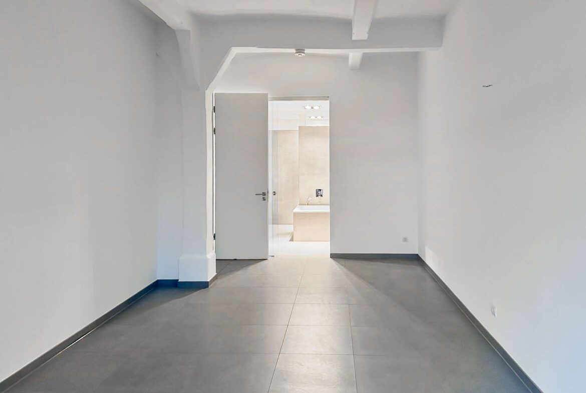 Long, empty hallway with light gray walls and darker gray floor tiles; white door at the far end is ajar, revealing a bright bathroom with a beige vanity.
