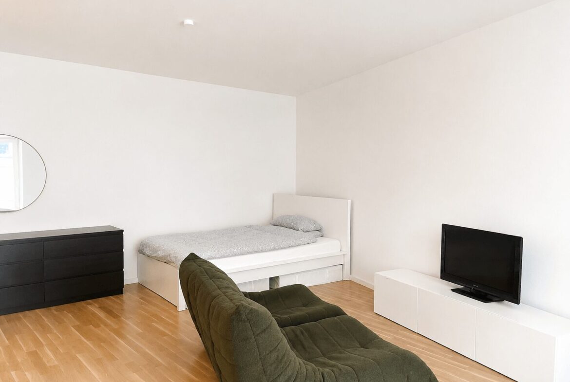 Minimalist bedroom with white walls; single bed, gray bedding, dark dresser with round mirror, olive-green lounge chair, and a low white TV stand with a TV on it.