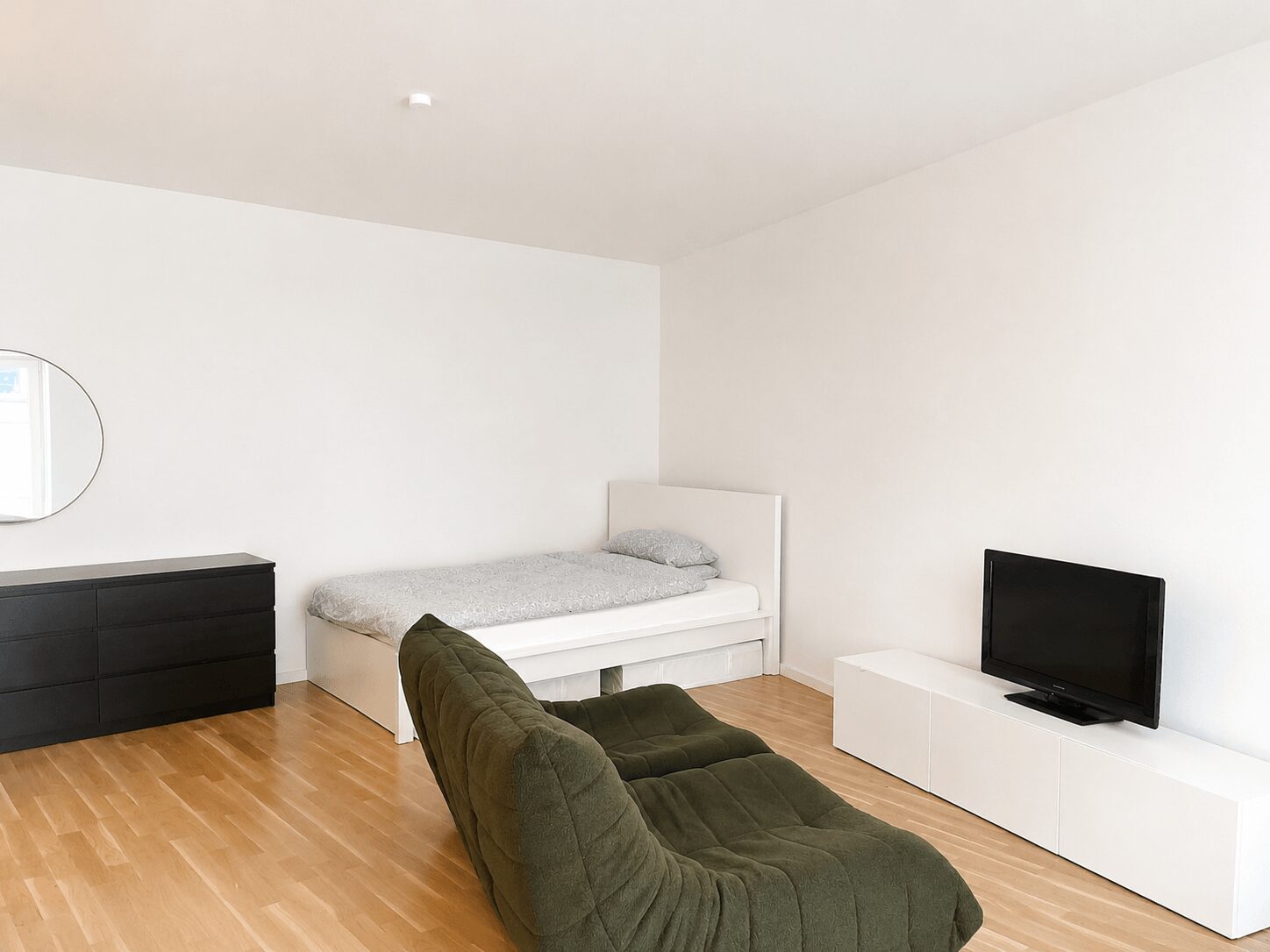 Minimalist bedroom with white walls; single bed, gray bedding, dark dresser with round mirror, olive-green lounge chair, and a low white TV stand with a TV on it.