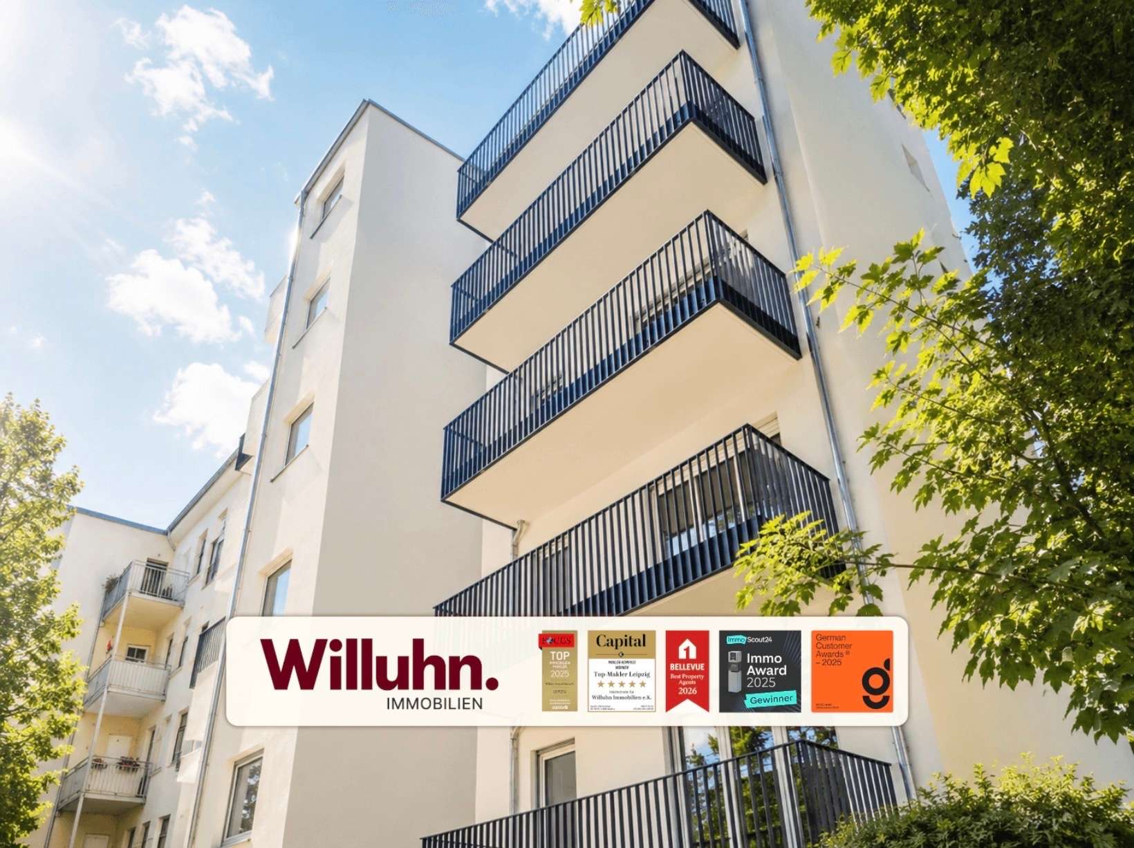 Modern white apartment building with stacked blue railings and balconies, sunlit sky and green trees on the right; foreground shows a Willuhn Immobilien banner.