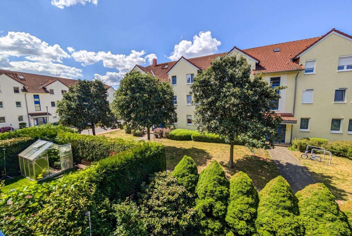 Sunny residential courtyard with trimmed hedges, trees, and pale yellow apartment buildings under a blue sky with white clouds, a glass greenhouse in the corner.
