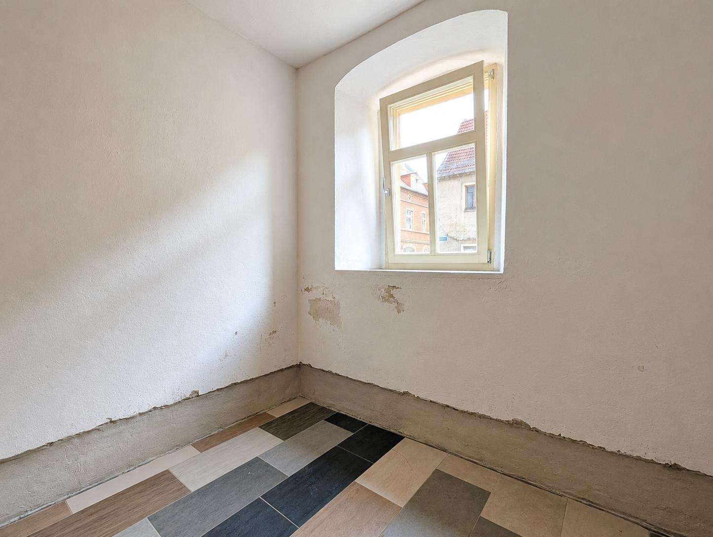 Corner of a bare room with peeling plaster, a small arched window letting in light, and a patterned tile floor in neutral tones.