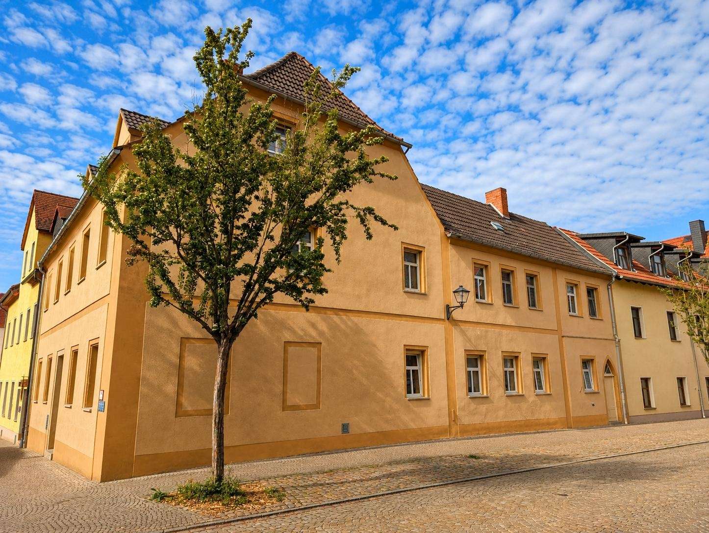 Peach-colored two-story building on a cobblestone street with a tree in front and a blue sky with clouds above.