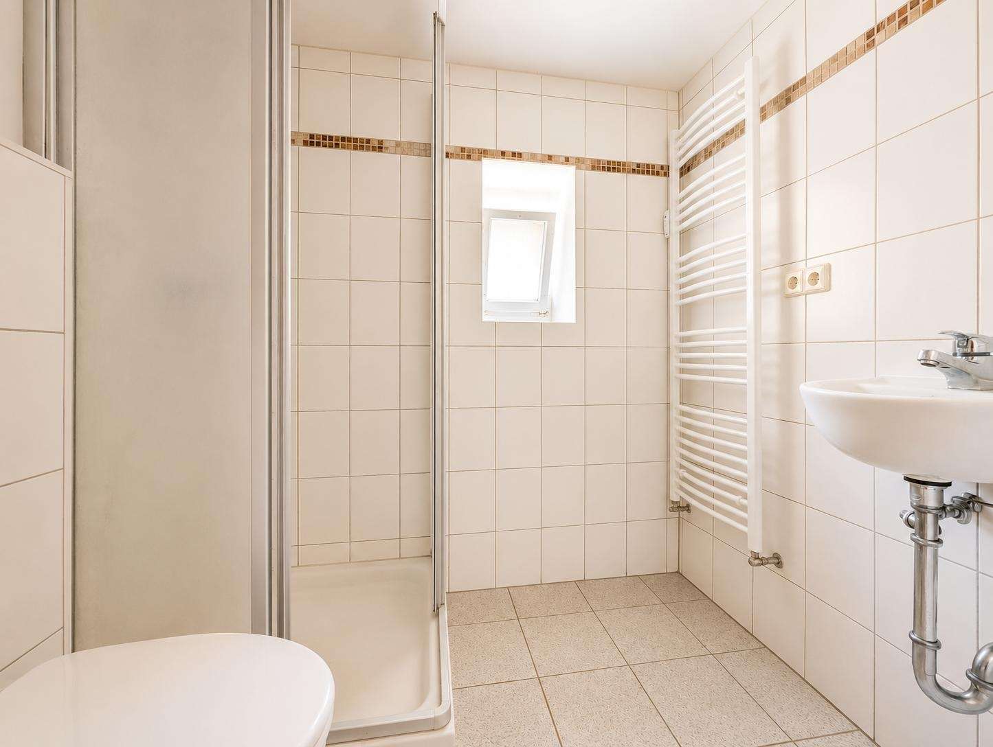 White-tiled bathroom with a corner shower, a white toilet in the foreground, and a wall-mounted towel radiator beside a sink on the right.