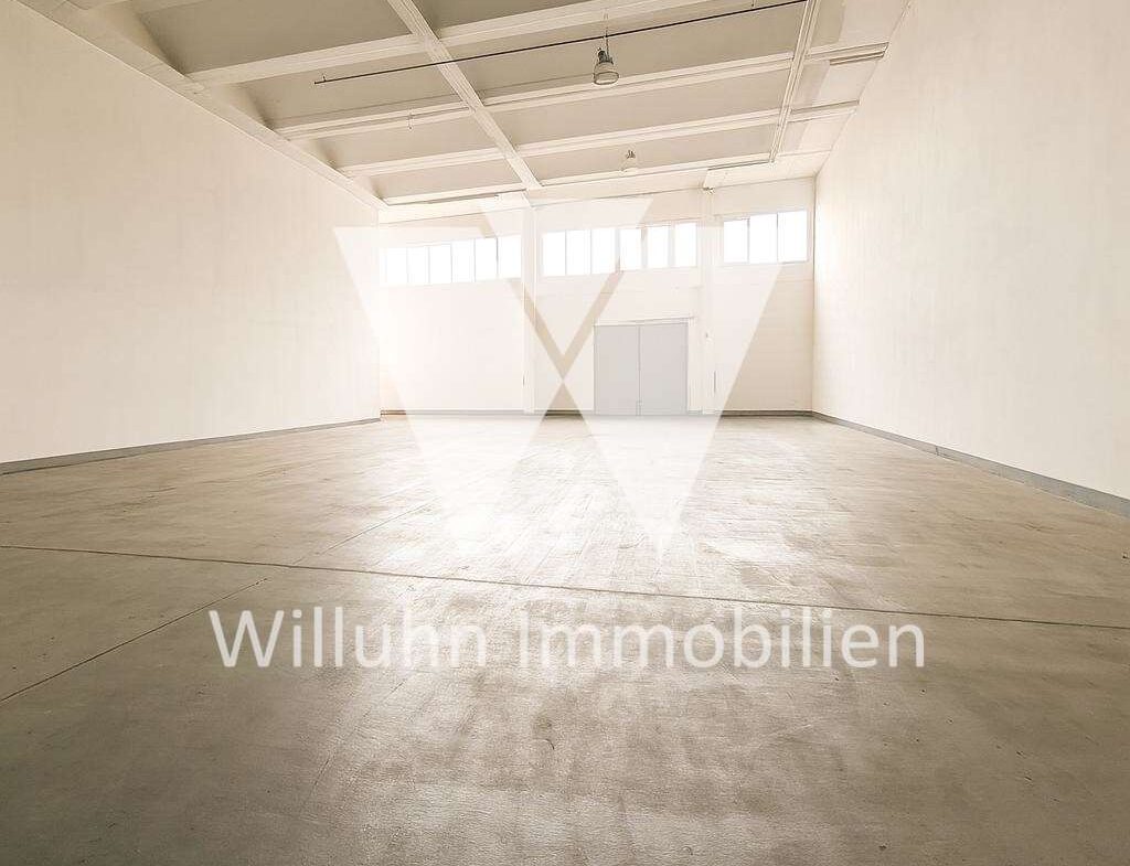 Empty white-walled warehouse with a high ceiling, exposed beams, and large windows at the far end. Watermark: Willuhn Immobilien.