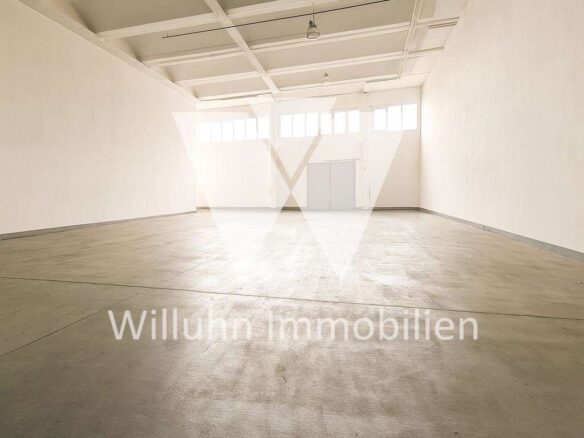 Empty white-walled warehouse with a high ceiling, exposed beams, and large windows at the far end. Watermark: Willuhn Immobilien.