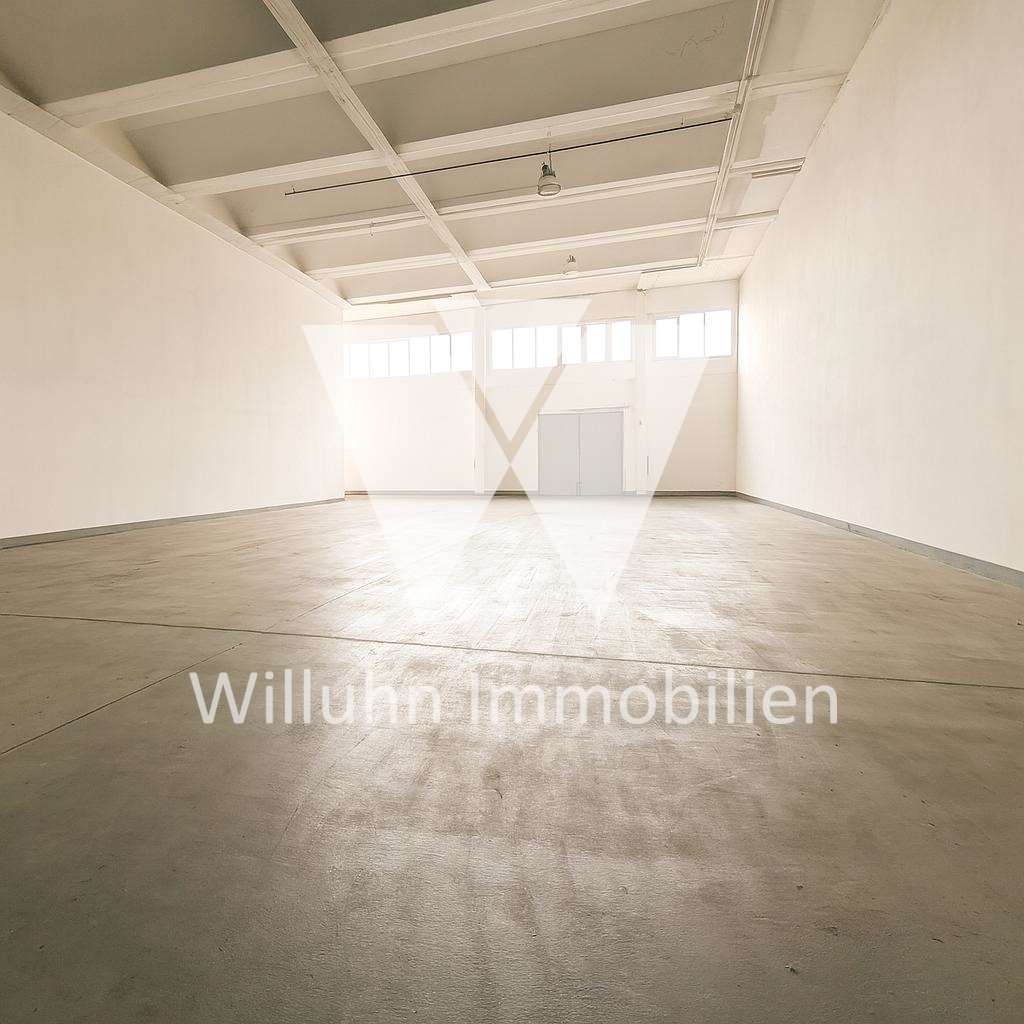 Empty white-walled warehouse with a high ceiling, exposed beams, and large windows at the far end. Watermark: Willuhn Immobilien.