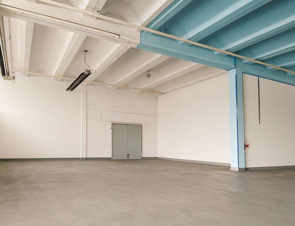 Empty industrial warehouse interior with white walls, gray double doors at the far wall, and blue steel beams supporting the ceiling.