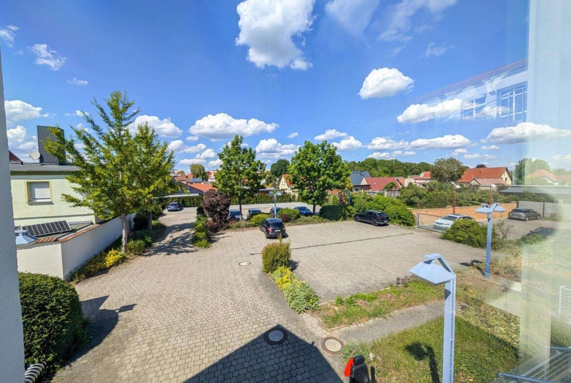View from a window over a sunny residential parking area with trees and houses against a blue, cloud-filled sky