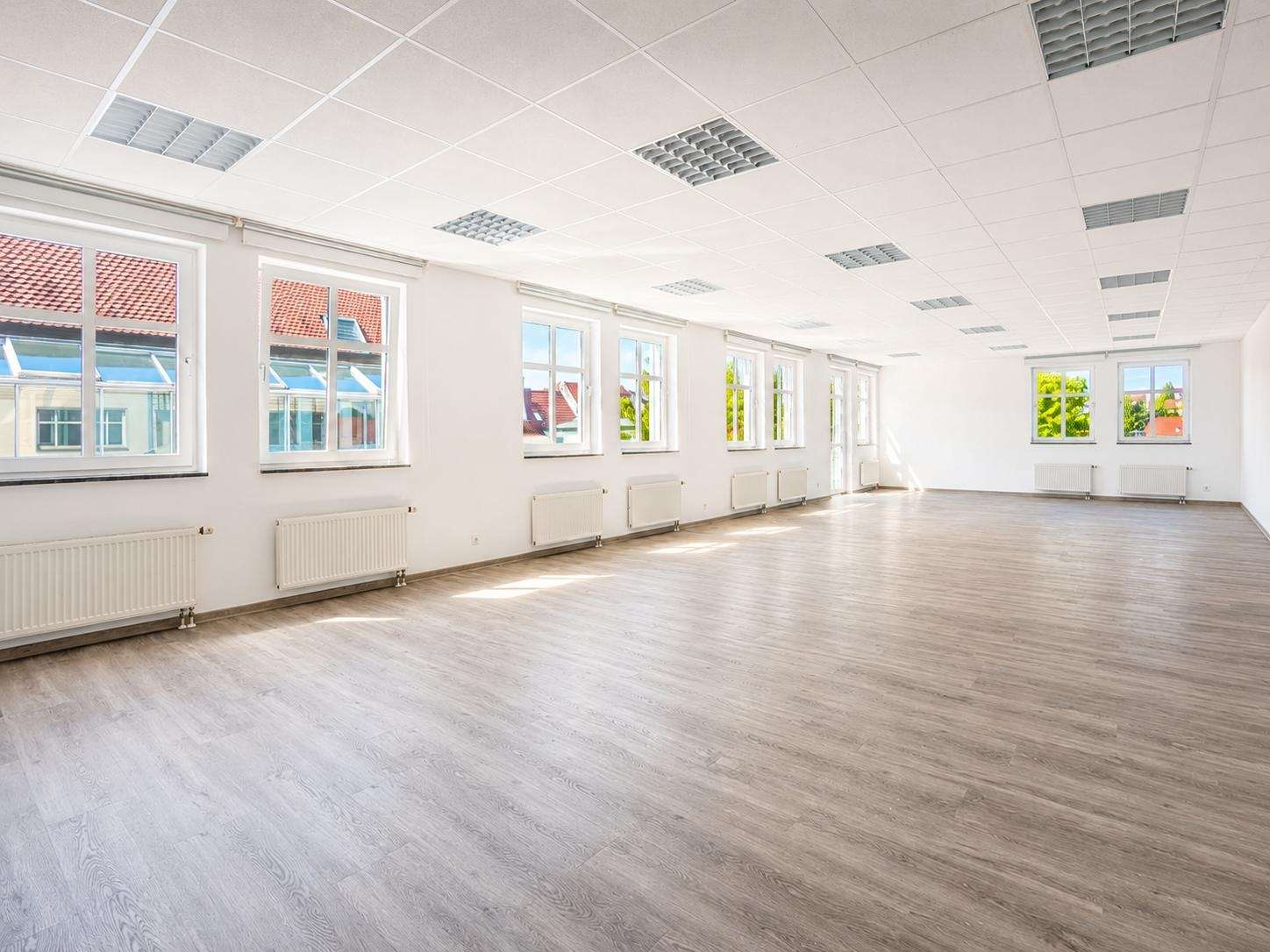 Bright empty multipurpose room with large windows along one wall, white walls, radiators, and light wood flooring, bathed in daylight outside.