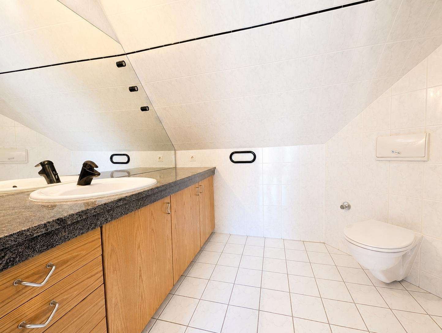 Bathroom with a sloped ceiling, double sink vanity with granite countertop, and wooden cabinets beneath. A wall-mounted toilet sits to the right, with black grab bars on the tiled wall nearby. White tiled floors and walls complete the scene.