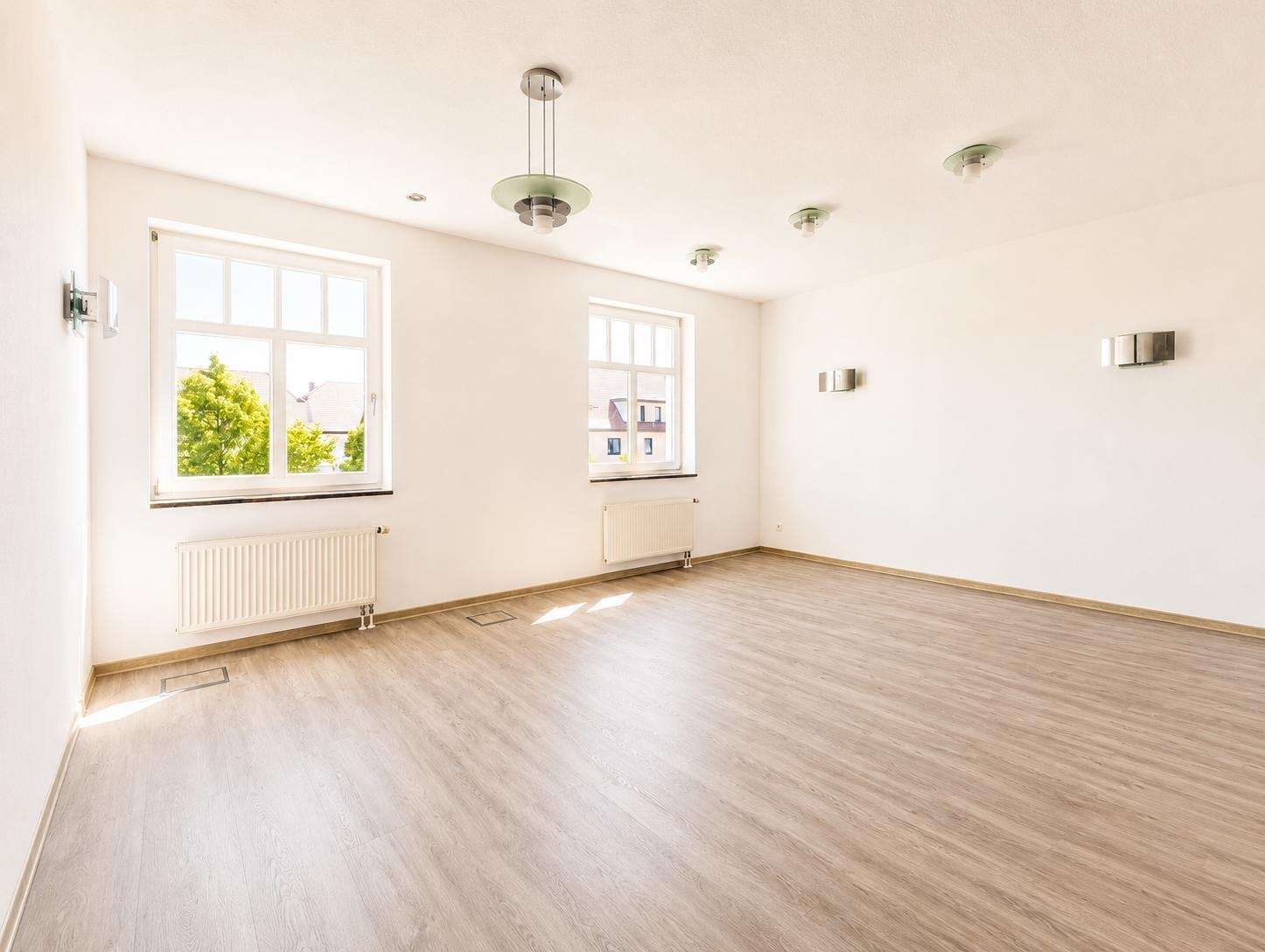 Bright, empty living room with light wood flooring, white walls, and two large windows letting in daylight; radiators beneath windows and modern ceiling lights.