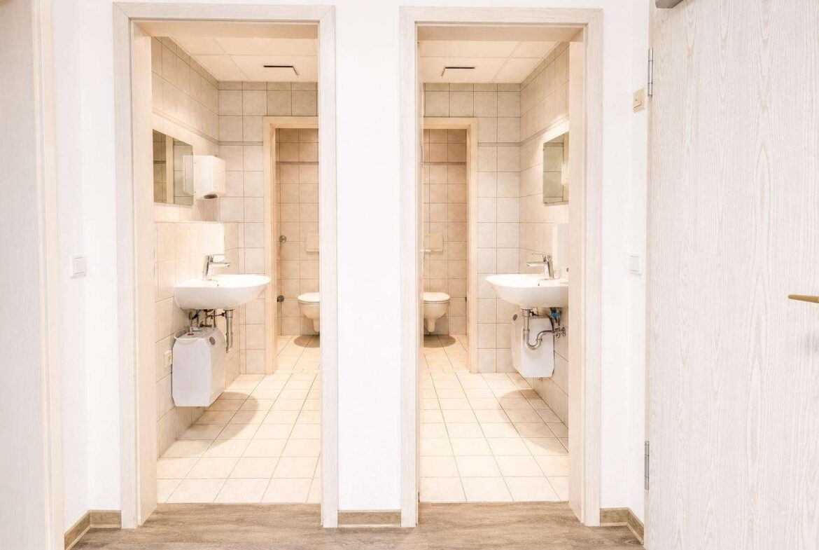 Bright hallway leading to two small tiled bathrooms with sinks and toilets visible through open doors; clean, neutral palette with beige tiles and white fixtures.