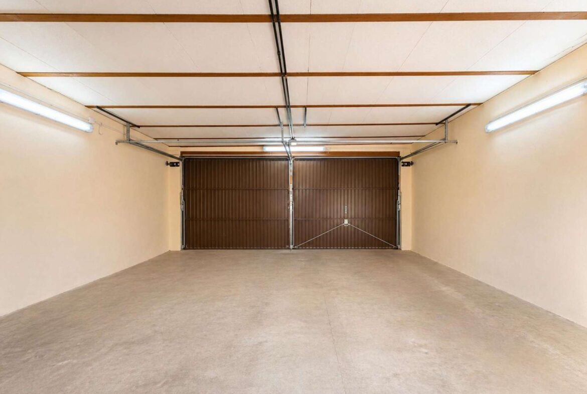 Empty indoor garage with brown sectional doors at the far end and bright fluorescent lighting along the walls.