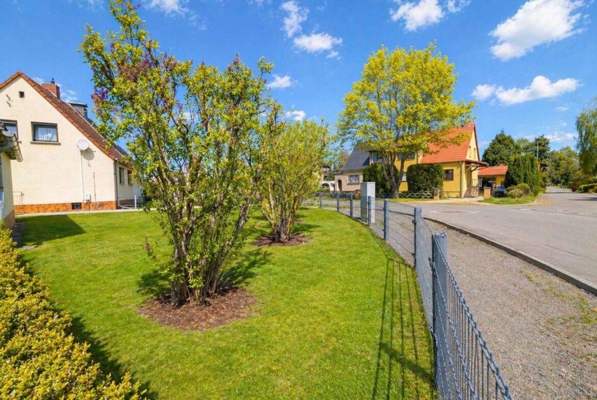 Suburban street scene with green lawns, trees, and yellow houses under a bright blue sky.