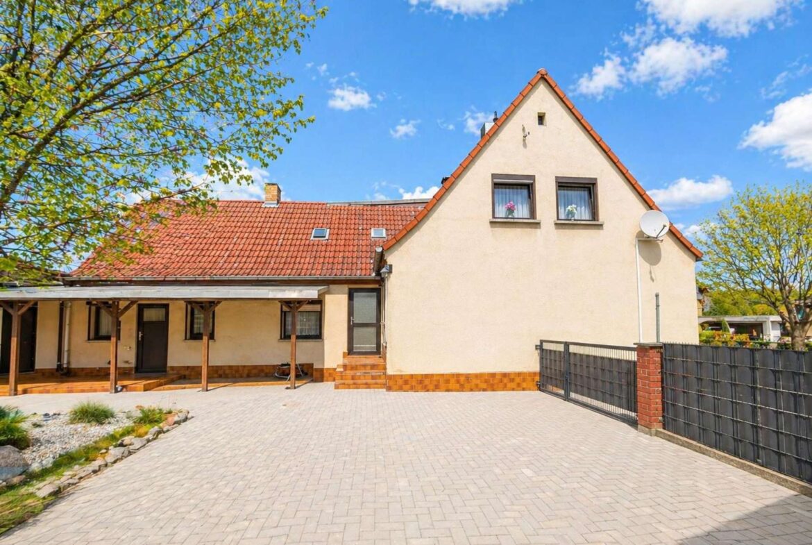 Housefront with red-tiled roof, beige walls, and two upper windows under a blue sky; a shaded porch is on the left.
