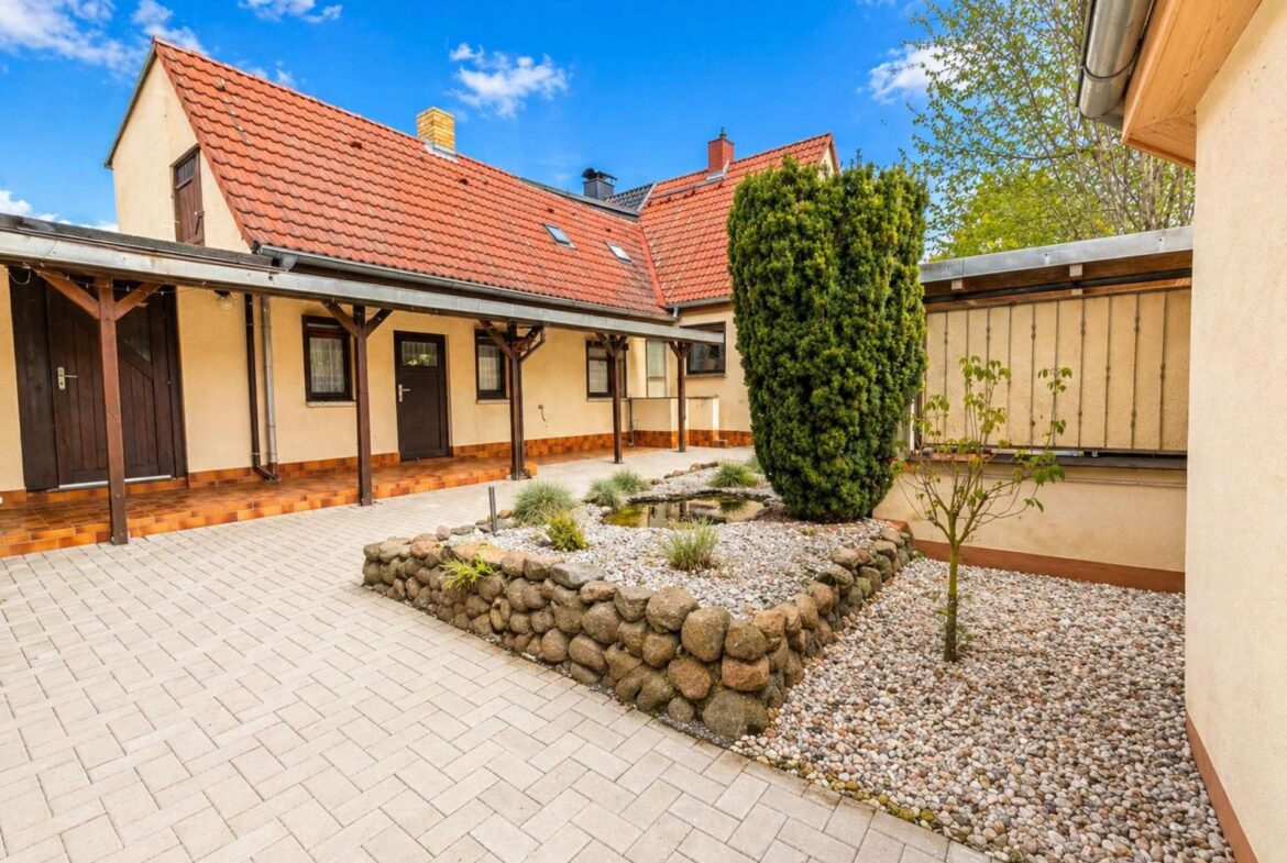 Courtyard of beige buildings with red-tiled roofs, wooden awnings, and a layered rock garden under a blue sky.
