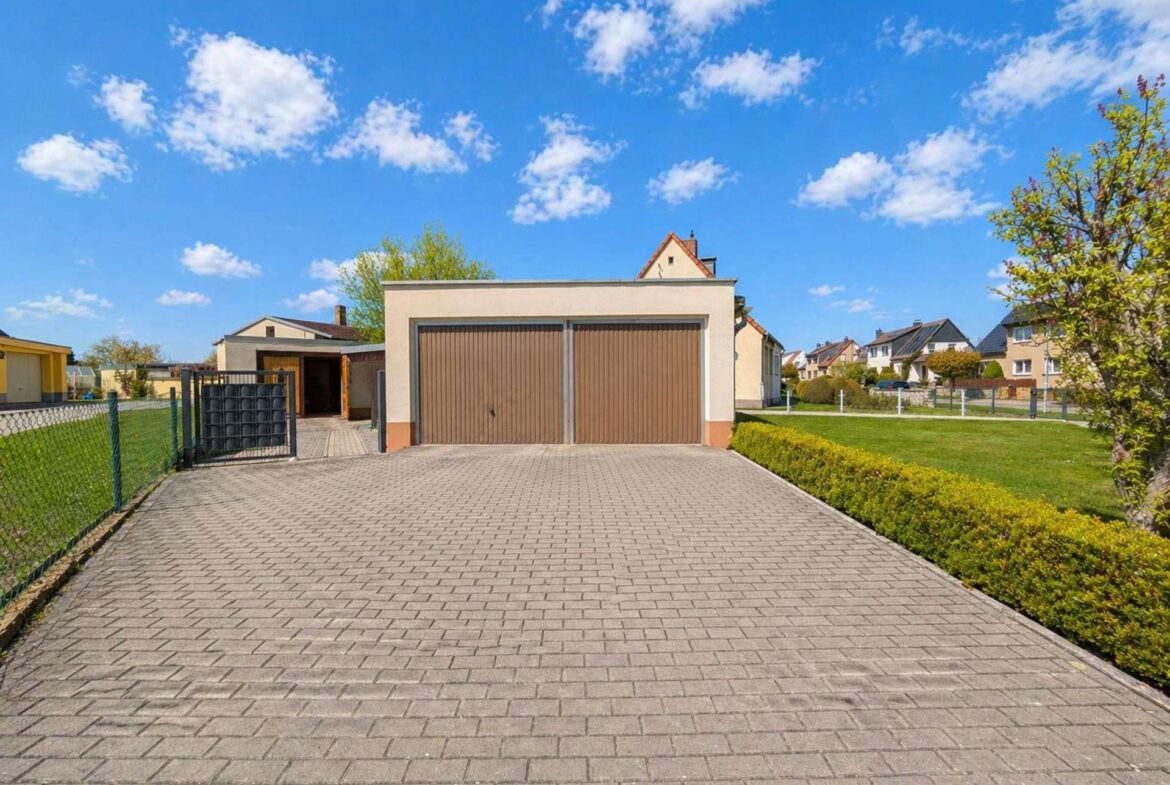 Front view of a beige house with a double brown garage, a long paved driveway, and green hedges in a suburban setting under a blue sky with scattered clouds.