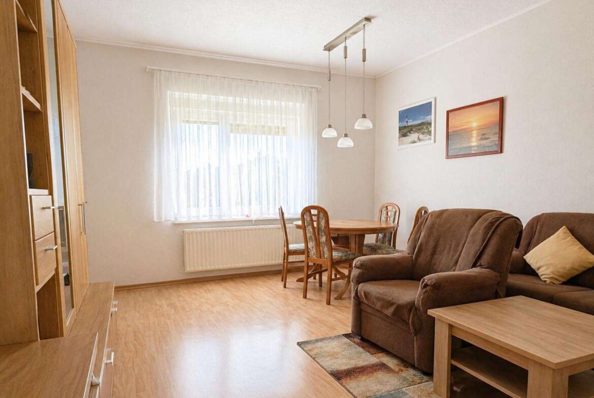 Cozy living-dining room with brown sofa, wooden coffee table, and a round dining table by a window with sheer white curtains; two framed beach photos on the wall.